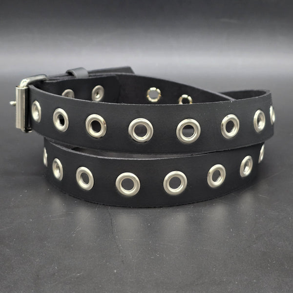 A black leather belt with a row of silver metal grommets running the length of the belt, coiled on a dark gray textured surface shown from the back