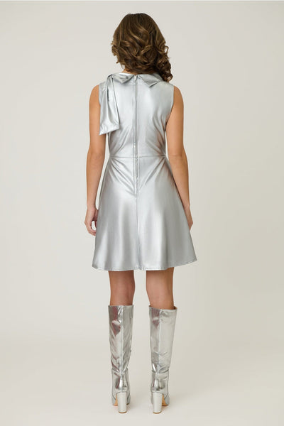 Model wearing a metallic silver knit fit and flare mini dress with cowl collar neck with asymmetrical self tie detail, darting at bust, and above the knee length skirt with pockets. Shown from back
