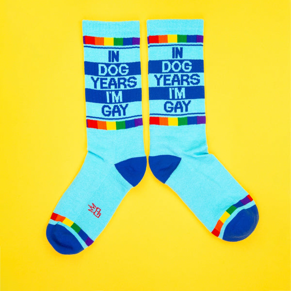 bright blue unisex crew socks with dark blue toes and heels & rainbow cuffs with "IN DOG YEARS I'M GAY" message on side. Shown on yellow background