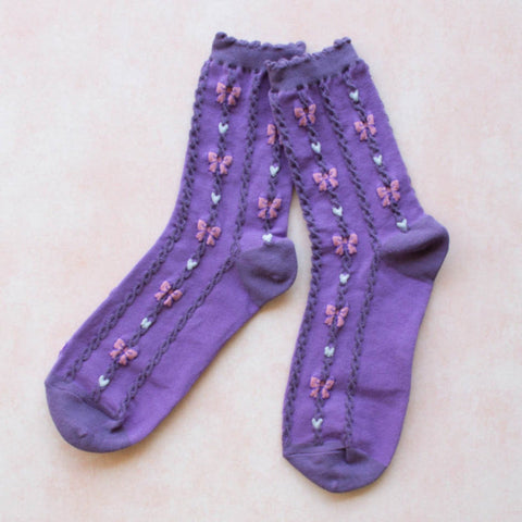 Purple socks with a ruffled cuff and an all over knit in pattern of white hearts and pink bows. Shown flat