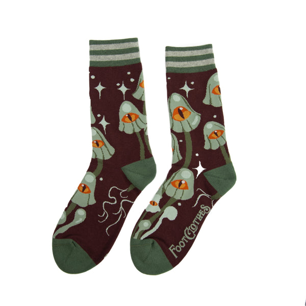 Unisex crew socks with an all-over pattern of green-grey mushrooms each with a large yellow and orange eye on their caps. On a purple-ish brown background surrounded by matching grey starbursts. Striped green and grey cuffs and solid green toes and heels. Seen flat from opposite side 