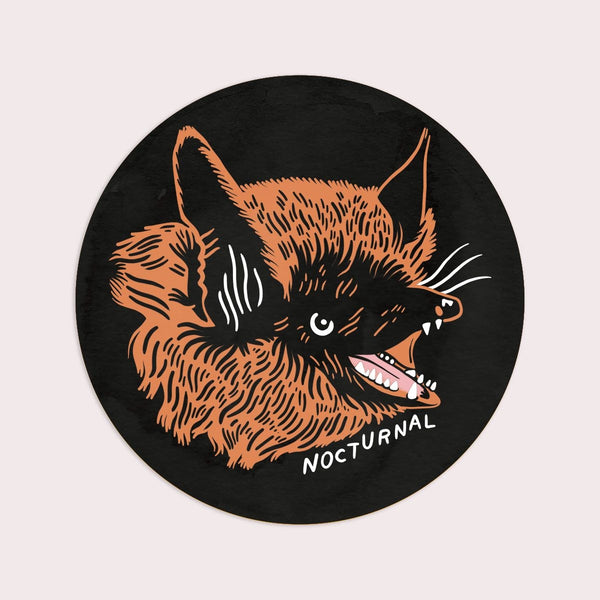 Round die-cut vinyl sticker of a brown bat head with open mouth and caption “NOCTURNAL” in white on black background