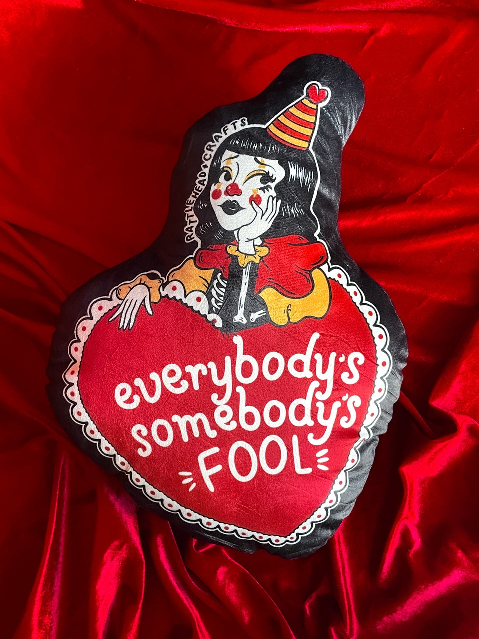 heart shaped plush pillow decorated with a sad yellow, red, and black outfitted clown girl and the message "everybody's somebody's fool" in the middle of the vintage valentine style heart with black background. Shown on red velvet background.