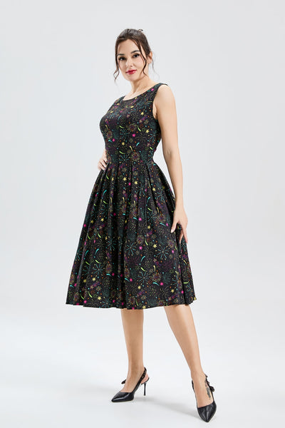 Model wearing a fit and flare dress made of soy silk in a black background pattern of exploding fireworks in bright red, pink, green, blue, orange, and purple. It has a high fitted neckline, princess seamed bodice, wide banded waist, and box pleated knee length skirt with pockets. Shown from a three quarter angle