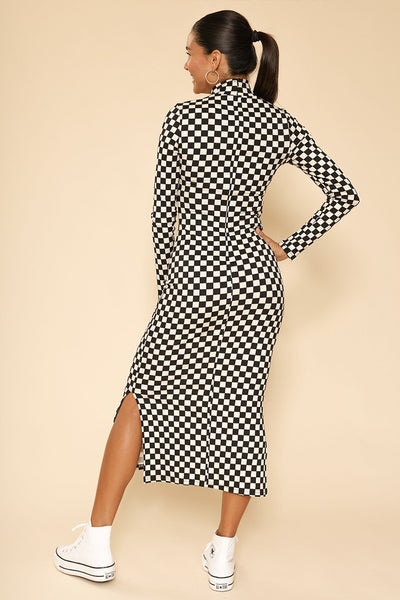 A model wearing a black and creamy white checkerboard pattern ribbed knit dress with long sleeves and a mock neck. Ends below the knee and has doubler slide slits. Shown from back