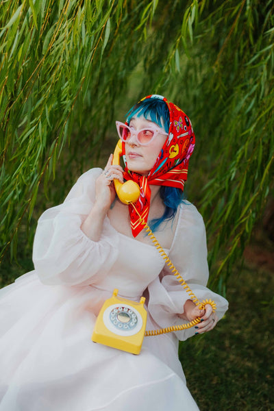 square satin twill scarf in bright red with yellow stripe border and all-over cartoon pattern of "junk drawer" items including bobby pins, heart locks, keys, dolphins, glue guns, shrimp, tiny cat-eye sunglasses, matches, buttons, and cigarettes. Shown worn wrapped around model's head