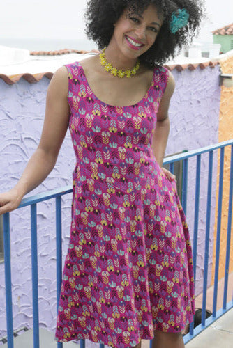 model wearing a stretch cotton sleeveless dress in a teal, cornflower blue, black, yellow, cream, purple and off-white repeat pattern of daisies and their stems in a chevron-like stripe pattern. The dress has a scoop neckline, reverse yoke waist, patch pockets, and full just below the knee length skirt