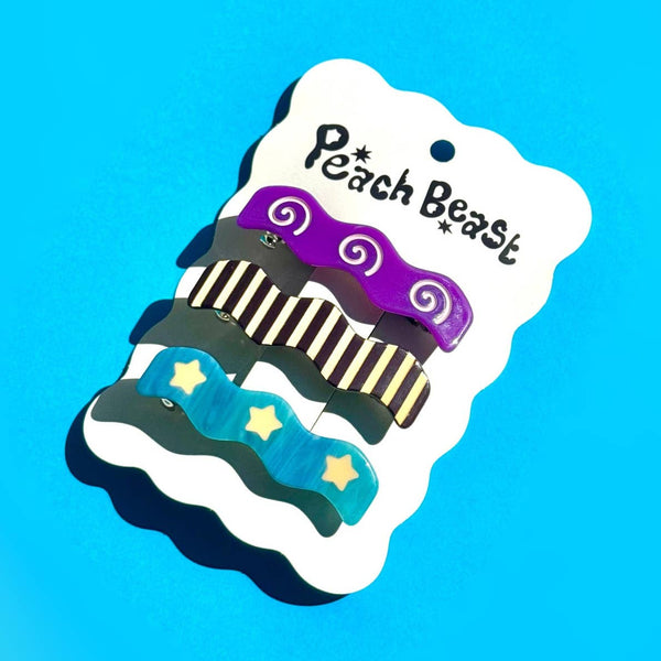 three acetate barrettes in a wavy rectangular shape with unique patterns (lavender spirals on purple background, black and cream stripes, pale yellow stars on marbled blue background ) attached to white backing card