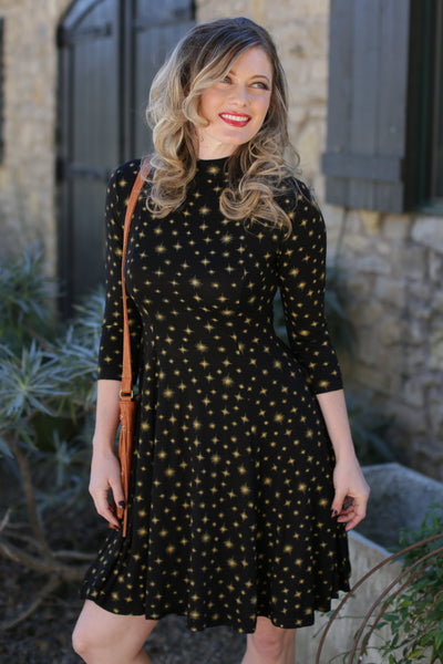 model wearing a stretch cotton knit long sleeved black dress with allover printed delicate gold starburst pattern that features a sleek mock turtleneck collar, 3/4 sleeves, flattering vertical seams for shaping, and finishes with a swingy just-above-the-knee length skirt