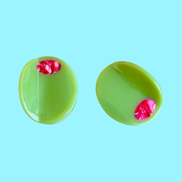 Two light green acetate mini hair clips in the shape of an olive with red marbled detail as a “pimento” middle. Shown on light blue background