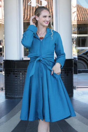 Model wearing capri blue fleece knee length coat with hood, zip front, matching waist tie, and full circle “skirt” 