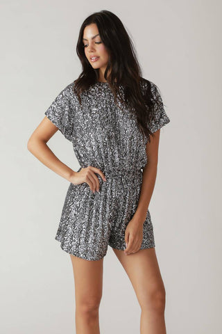 model wearing a short sleeved romper in silver sequin fabric with zigzag stripe pattern, round neckline, gathered elasticized waist, and deep keyhole detail at back. Shown from front