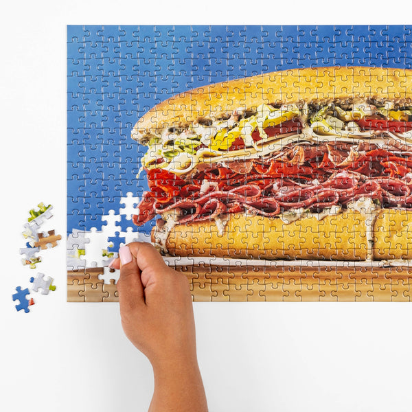 Jigsaw puzzle of a sub sandwich with a hand holding a piece, on a white background