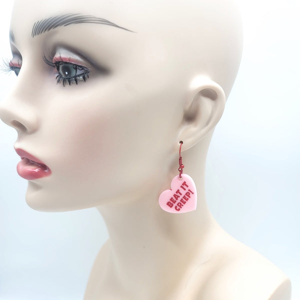 pink laser cut acrylic heart shaped dangle earrings resembling conversation heart candies with the message "BEAT IT CREEP!" in red capital letters. Shown on mannequin head