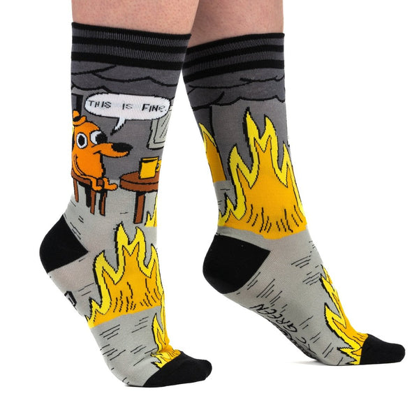 black, grey, yellow, and orange unisex crew socks with black and grey stripe cuffs and black toes/heels with all over pattern of a cartoon dog wearing a hat sitting at a kitchen table surrounded by flames with a speech bubble saying "THIS IS FINE." Shown worn from the side