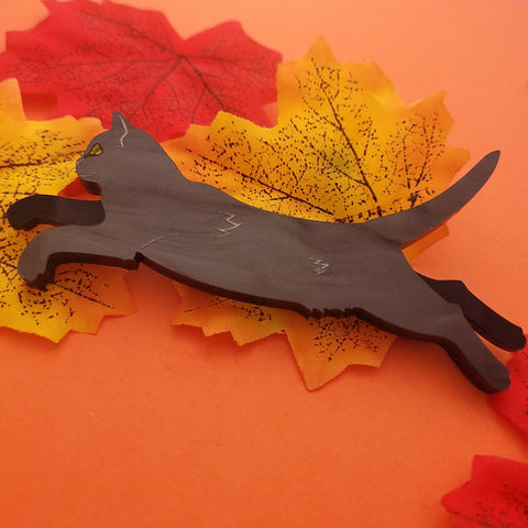 A laser-cut acrylic brooch in a rippled black pattern of a black cat with painted silver detailing. Cat is pictured with limbs out and body extended. Yellow jewel detail for eye. Shown flat on an orange autumn leaf background 
