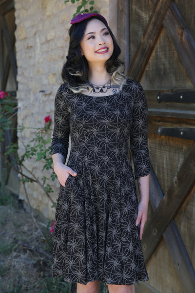 Model wearing a black stretch cotton knit dress with allover printed pattern of cream pointillism spiderweb. Dress has open neckline, three quarter sleeves, side seam pockets, vertical seaming, and a knee length full skirt 