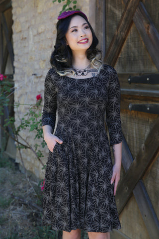 Model wearing a black stretch cotton knit dress with allover printed pattern of cream pointillism spiderweb. Dress has open neckline, three quarter sleeves, side seam pockets, vertical seaming, and a knee length full skirt 