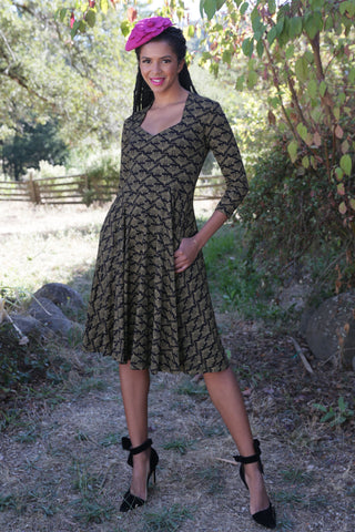 model wearing knit cotton dress in black with gold printed stylized floral pattern. Dress has Queen Anne neckline, three quarter sleeves, full skirt and pockets