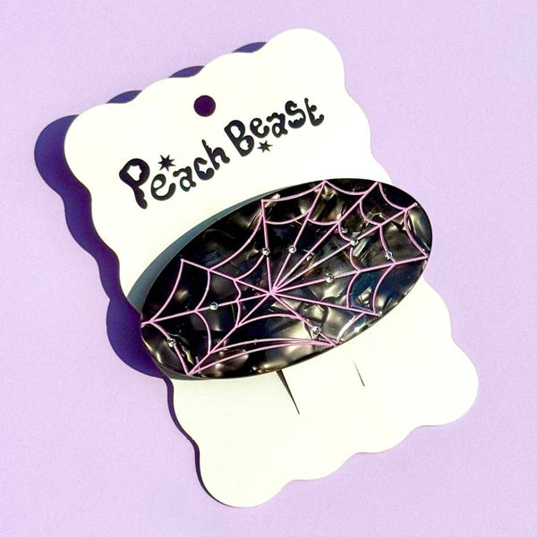 A black oval hair claw clip made of marbled black plant-based cellulose with a pink spiderweb design and small silver rhinestones, displayed on a white card labeled 'Peach Beast' against a light purple background.