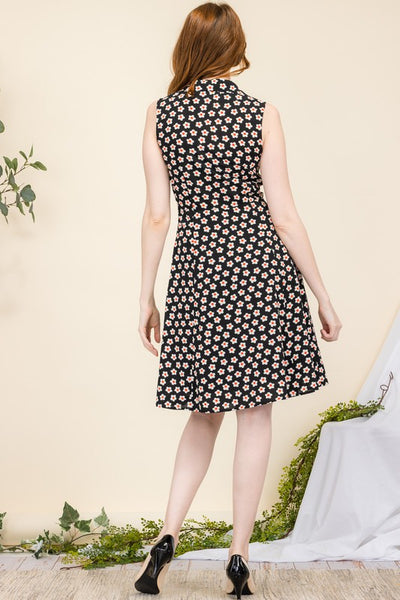 model wearing a sleeveless shirtwaist dress in a black background white and red daisy pattern. It has red plastic buttons running down the bodice and a v-neck shawl style collar. It has a slightly flared skirt ending at the knee. Shown from the back