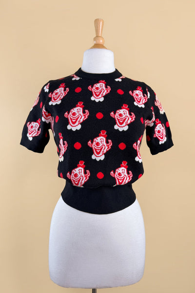 short sleeve acrylic knit sweater with black background and all-over tessellated white and red clown head print alternated with red polka dots. The top has a crew neck and ribbed hem. Shown from front