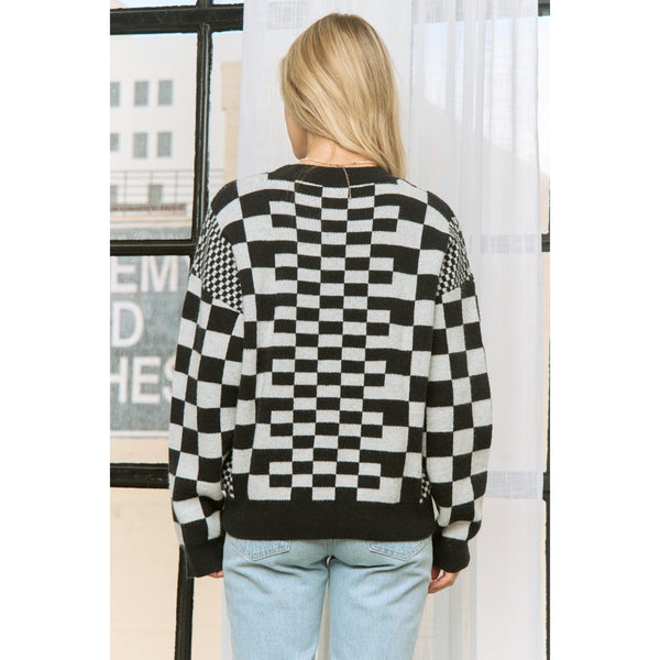 Woman wearing a black and white checkered sweater with various sizes of checker patterns and ribbed black collar, cuffs, and bottom hem. Shown from back