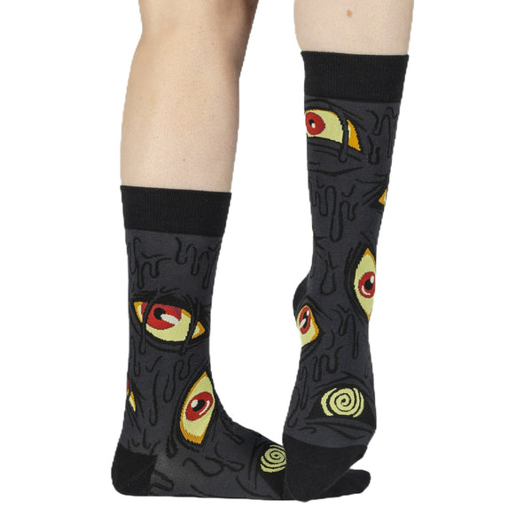 A pair of unisex cotton blend greyish black background crew socks with a repeating pattern of stylized eyes in yellow with red pupils and dripping oozing shapes. Shown worn by model from front to display swirled eyeballs on top of foot