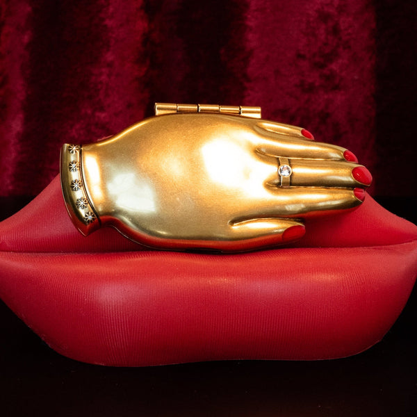 bright slightly burnished finish gold hand-shaped compact mirror with red enameled manicure and clear jeweled ring and bracelet detail. shown closed on red lip-shaped surace with red velvet background