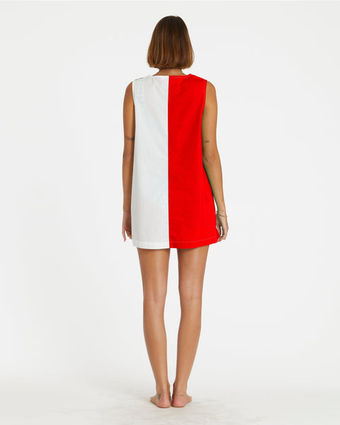 a model wearing a cotton a-line smock style mini dress in half red, half white design divided vertically down the middle of the garment. it has a round neckline with slash detail, button closure running down the left shoulder and down the side of the dress, and patch pockets in alternate colors at each hip. Shown from back