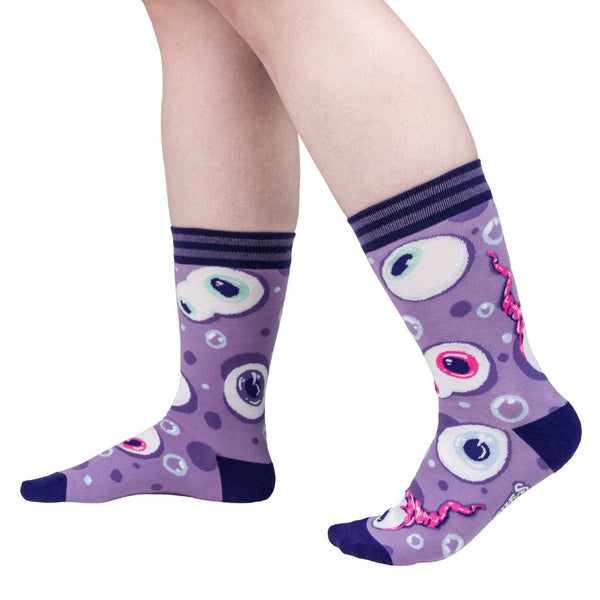 cotton unisex crew socks with allover pattern of light purple background and eyeballs with pink, purple, or blue irises. Some eyes have pink optic nerves. Surrounded by blue bubbles. Dark purple striped cuffs and solid purple toes and heels. Shown worn by a model from the side