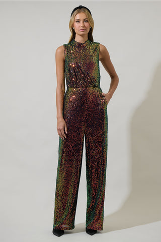 model wearing an iridescent sequin jumpsuit with shades of green, yellow, orange, and purple. Jumpsuit is sleeveless, has a mock neck, asymmetrical draped bodice detail and wide leg pants. Shown from front