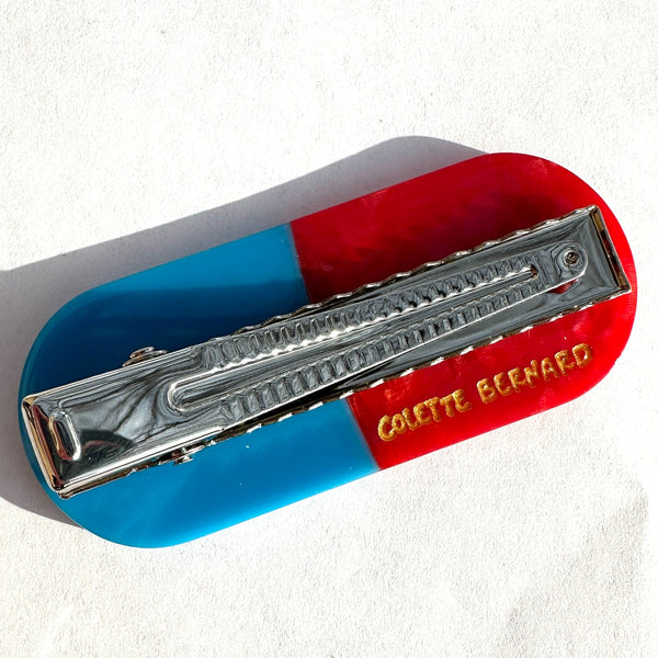 bioacetate hair clip in the shape of a solid light blue and marbled red pill capsule. Shown from back on white background to display alligator style silver metal clip and gold branding