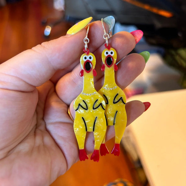 yellow marbled acetate dangle earrings in the shape of a pair of rubber chickens with red mouths, feet, and waddles with silver rhinestone jeweled necklaces. Shown held by hand