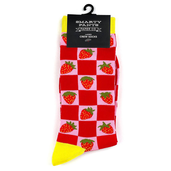 Crew socks with a knit-in pattern of strawberries on a pink and red checkered background with bright yellow cuffs, toes, and heels. Shown flat in packaging from front 