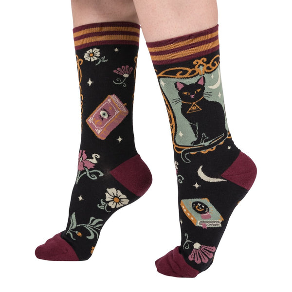 Cotton blend unisex crew socks with framed illustration of black cat on mint green background with black background pattern of flowers, stars, moons, and spell books. Socks have maroon toes and heels & maroon and golden yellow striped cuffs. Shown worn by model from side