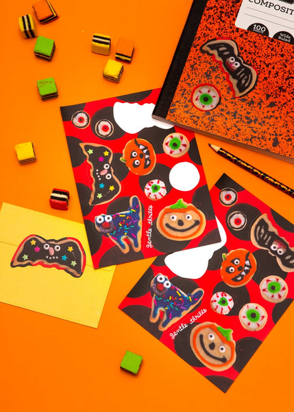 sticker sheet of Halloween-themed cookies with colorful frosting designs on a red and black polka dot background. Shown on orange background with stickers applied to notebook and surrounded by candy