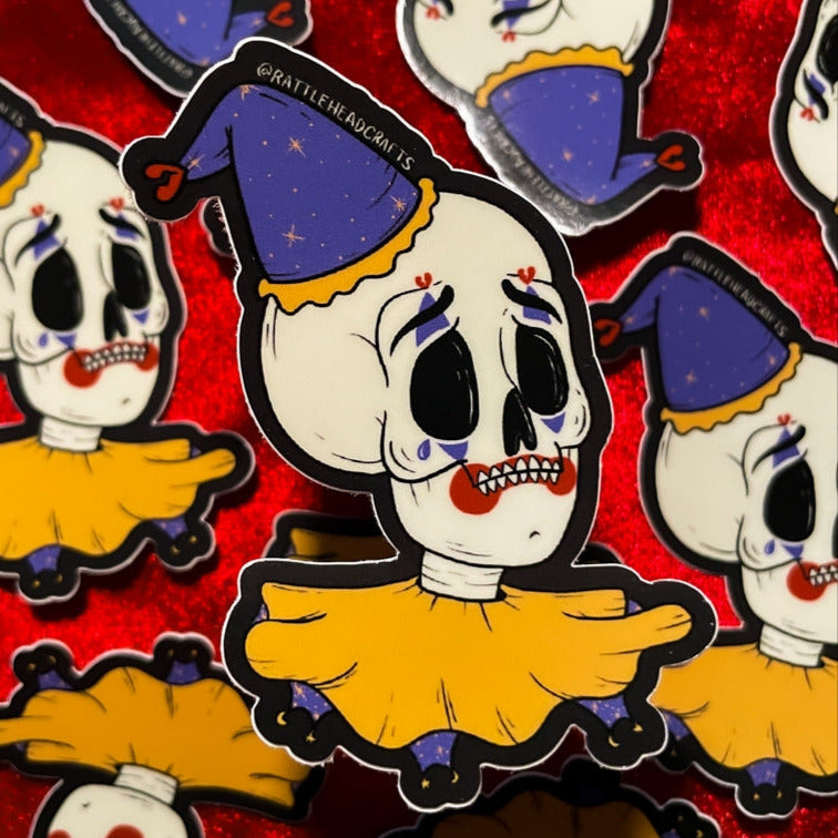 glossy finish die-cut vinyl sticker featuring an illustration of a crying skeleton with red and purple clown makeup sporting an orange and purple Pierrot style hat and ruffled collar
