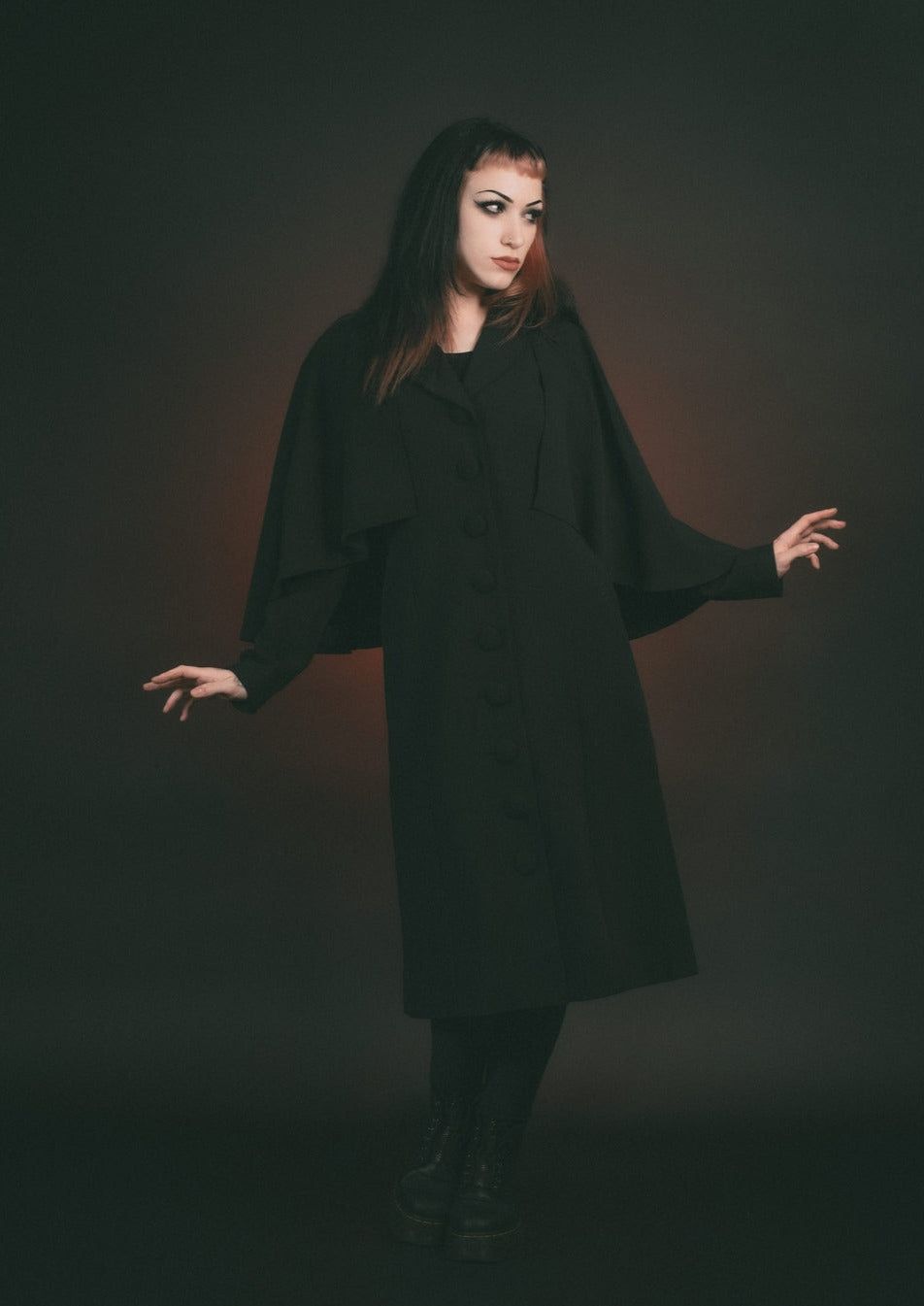 Model wearing black full length faux wool-blend coat with shawl collar and attached elbow-length cape. Shown with arms outstretched
