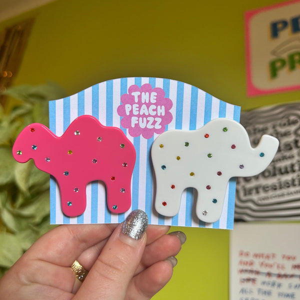 pair of acetate barrettes in the shape of a pink camel and a white elephant animal cracker, both decorated with multicolor jewels as sprinkles. Shown on striped backing card