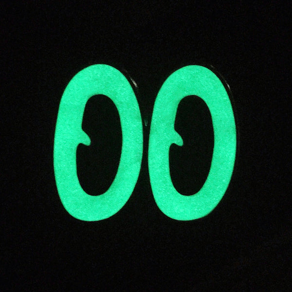 gold metal enamel pin of a large pair of oval-shaped cartoon eyes with black irises and bright white whites. Shown flat in the dark to display glow-in-the-dark capabilities 
