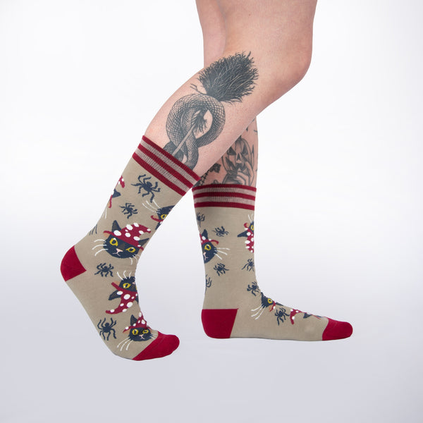 Unisex crew socks in light brown with red striped cuffs and red toes & heels. All over pattern of black cats wearing red and white dotted witch hats and small black spiders. Shown worn by model 
