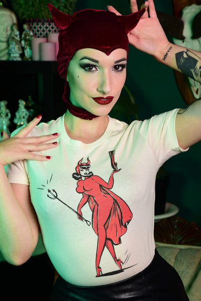 model wearing ivory t-shirt with printed illustration of vintage style pinup wearing devil costume with horns and cape, holding a black trident in one hand and a fetish-style high heeled boot in the other
