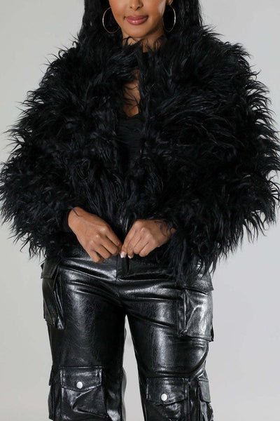 A model wearing a voluminous black shaggy faux-fur cropped open front jacket over a black top and shiny black faux-leather cargo pants. Shown from front