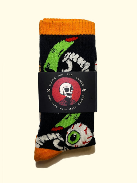 black cotton crew socks with orange cuffs, heels, and toes with an all-over pattern of plastic vampire teeth, witch fingers, and green bloodshot eyeballs. Shown flat in packaging