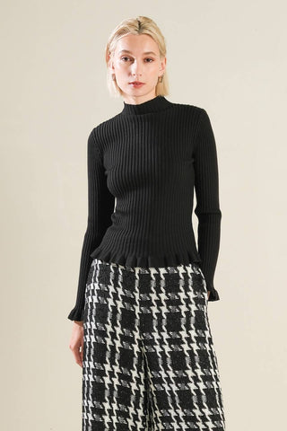 model wearing ribbed knit black mock turtleneck with long sleeves and ruffle detail at each cuff and bottom hem. Shown from the front