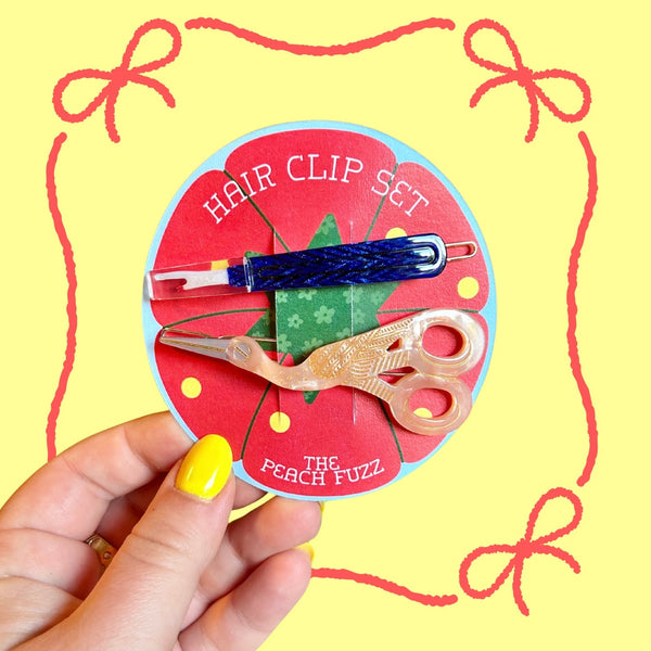 set of barrettes in the shape of stork scissors and seam ripper made of cellulose acetate shown on packaging on a yellow background