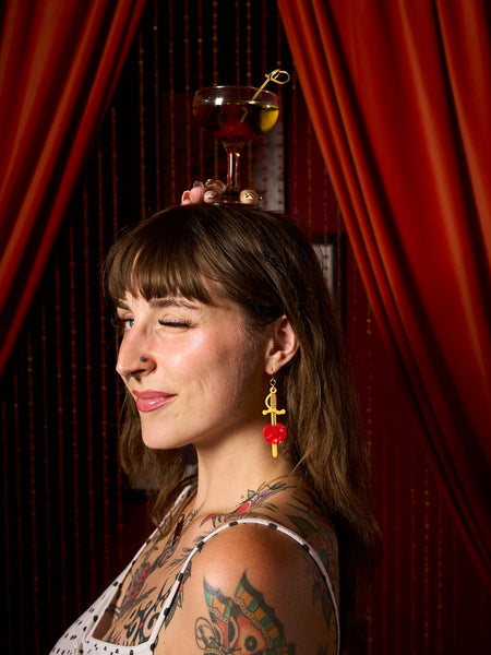 acetate dangle earrings in the shape of a pair of yellow cocktail sword skewers with a marbled maraschino cherry with silver rhinestone decoration. Shown worn by a model holding a drink on top of her head