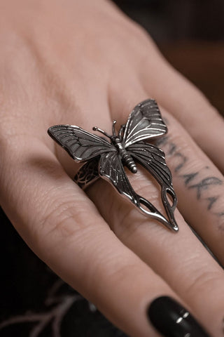 Model wearing a stainless steel ring in the shape of a highly detailed butterfly with long hind wings that meet just past the model’s knuckle. The butterfly is attached to an ornately detailed band. 