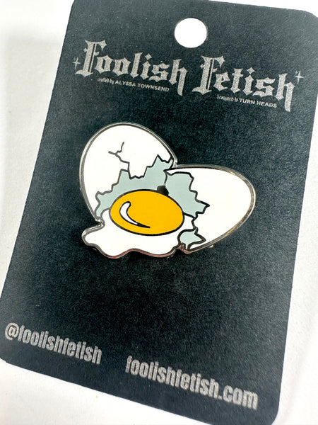 silver metal enamel pin of a white egg cracked in two pieces with a large yellow-yoked egg in between. Shown on black backing card